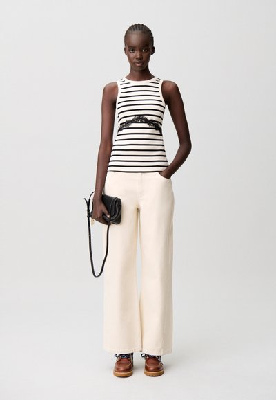 Top senza maniche a righe con dettaglio in pizzo, abbinato a pantaloni larghi crema e scarpe marroni con lacci. La modella tiene una borsa a tracolla nera.
