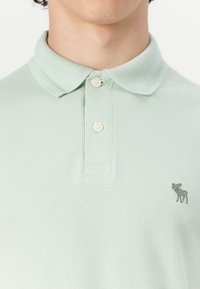 Light green polo shirt made of textured fabric with a collared neck, two buttons, and a small embroidered moose logo on the left chest.