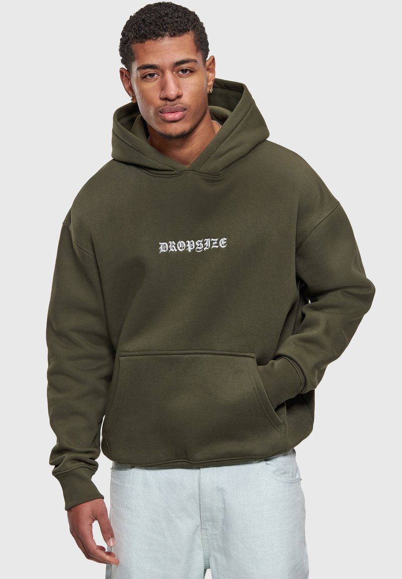Joven con sudadera con capucha de color verde oliva con el texto "DROPSIZE", una mano en el bolsillo y pantalones de color claro frente a un fondo liso.