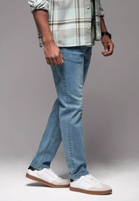 Jeans de mezclilla azul claro con corte slim, combinados con zapatillas blancas con detalles en negro y beige. El conjunto incluye una camisa de cuadros.