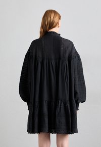 Črna obleka s plastehnim dizajnom, ki ima nabran material na rokavih, visok ovratnik in čipkast detajl ob telesu.