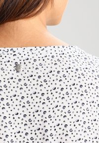 Tissu blanc à motif de points noirs, avec un col rond et une étiquette logo métallique à l'arrière. Texture douce et design léger.