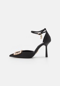 Scarpa con tacco alto nera a punta con cinturino alla caviglia, dettaglio con fibbia in oro e cristallo sulla punta e piccolo charm dorato sulla fibbia del cinturino.