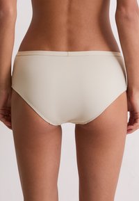 Frau in beiger nahtloser Unterwäsche, von hinten gezeigt, vor einem schlichten hellen Hintergrund.