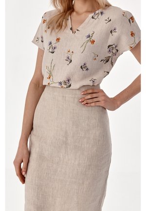 Vrouw draagt een beige korte mouwen blouse met bloemenpatroon, ingestopt in een beige linnen rok met hoge taille, hand op heup tegen een effen achtergrond.