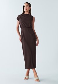 Robe longue marron texturée avec une épaule dénudée, taille cintrée et fente latérale. Associée à des sandales tressées de couleur claire.