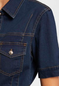Tummansininen denim-paita, jossa on lyhyet hihat, terävä kaulus ja rintatasku metallinappilla. Eristyssäikeet saumoissa.