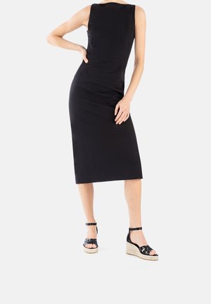 Schwarzes ärmelloses Kleid mit einer figurbetonten Silhouette, knielang, kombiniert mit schwarzen sandalenartigen Wedges mit einem strukturierten Espadrille-Sockel.
