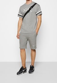 Muž v šedém tričku Nike a ladících šortkách, s černými teniskami Nike a černou krosnou, stojící na bílém pozadí.