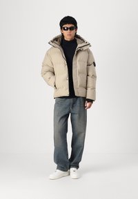 Beige pufferjack met een hoge kraag, ritssluiting aan de voorkant en knopen. Gecombineerd met donkere jeans en witte sneakers. Zwarte zonnebril maakt de look compleet.