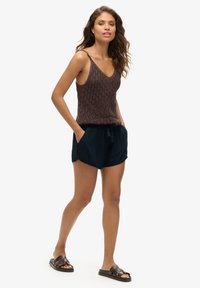 Débardeur en crochet marron à fines bretelles, associé à un short bleu foncé à taille élastique avec des poches latérales. Des sandales à glisser marron complètent l'ensemble.