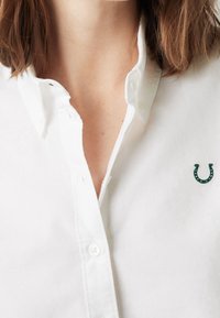 Chemise blanche à boutons avec col, ornée d'un petit logo fer à cheval vert sur le côté gauche. Texture de tissu lisse.