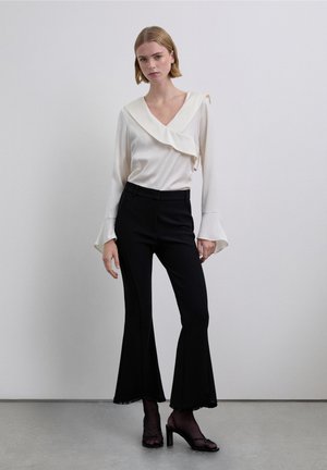 Blusa crema con volantes y mangas largas acampanadas, combinada con pantalones negros de campana con dobladillos deshilachados y tacones negros con tiras. Fondo sencillo.