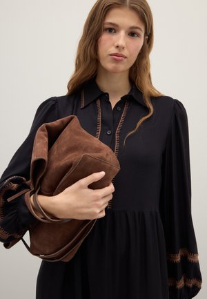 Schwarzes Kleid mit Puffärmeln und braunen Nähten entlang der Knöpfe; hält eine weiche braune Wildledertasche mit Ledergriffen.