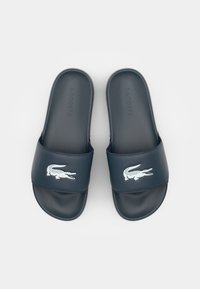 Navy blue slide sandals with a matte finish, featuring a white crocodile logo and embossed "LACOSTE" branding on the upper band.