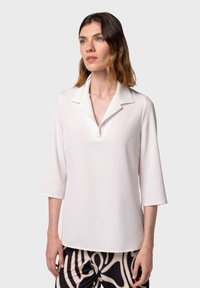 Polo shirt bianco a maniche lunghe con scollatura a V, caratterizzato da un colletto strutturato e una texture liscia. Indossato con pantaloni a righe bianchi e neri.