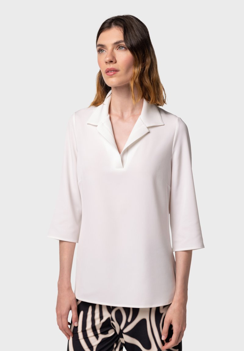 Polo shirt bianco a maniche lunghe con scollatura a V, caratterizzato da un colletto strutturato e una texture liscia. Indossato con pantaloni a righe bianchi e neri.