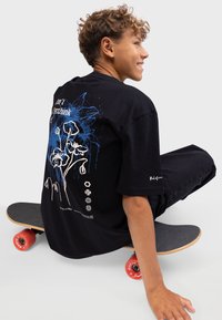 Schwarzes Baumwoll-T-Shirt mit blauem Blumenmotiv auf dem Rücken, mit dem Text "Don't Overthink." Das Model sitzt auf einem Skateboard mit roten Rädern.