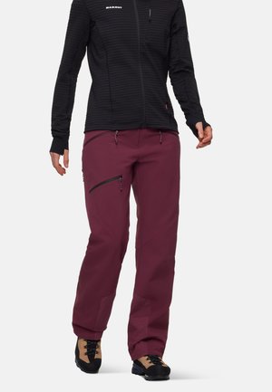 Persoon draagt een zwarte jas met ritssluiting en duimgaten en donkerpaarse broek met ritszakken, gecombineerd met beige en zwarte wandelschoenen.