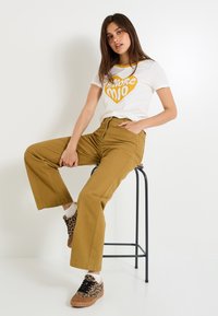 Camiseta blanca con gráfico de corazón amarillo, combinada con pantalones de pierna ancha mostaza y zapatillas marrones con estampado de leopardo. Fondo simple.