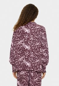 Donna con capelli ricci che indossa una blouse a maniche lunghe color bordeaux e rosa chiaro con un motivo astratto e pantaloni abbinati, vista da dietro.