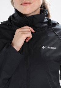 Osoba zapínající černou bundu Columbia, s viditelným logem značky na hrudi a vysokým límcem kolem krku.