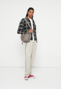 Borsa a tracolla grigia con cinturino regolabile, pantaloni khaki a vestibilità comoda, camicia a quadri nera e verde sopra una maglietta bianca e sneakers rosse.