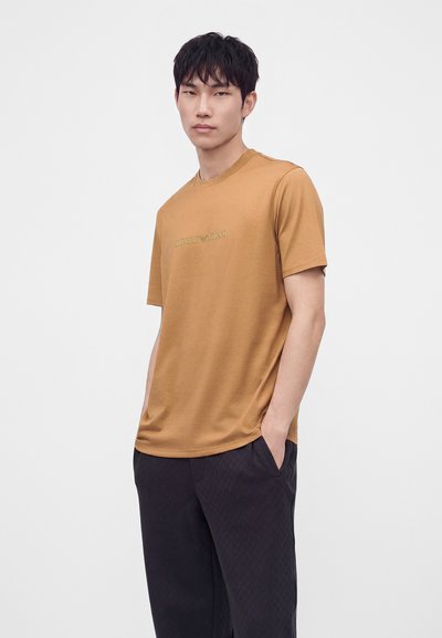 Άνδρας που φοράει μια μουσταρδί μπλούζα Emporio Armani και μαύρο παντελόνι, στέκεται με το ένα χέρι στην τσέπη του μπροστά σε απλό φόντο.
