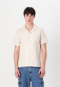 Kurze Ärmel, cremefarbenes Hemd mit einem dezenten kreisförmigen Muster, einer Tasche und Knöpfen, kombiniert mit blauen Jeans.