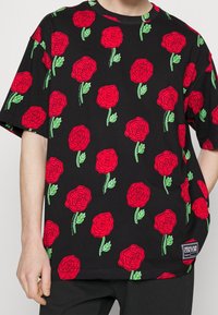 Svart t-shirt med stora röda rosor och gröna bladmönster. Lätt och lös passform med korta ärmar och en etikett i nedre vänstra hörnet.
