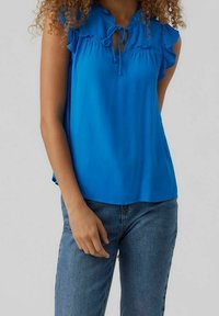 Donna che indossa una blusa senza maniche di un vivace blu con un colletto annodato e spalle a balze, abbinata a jeans di denim blu.