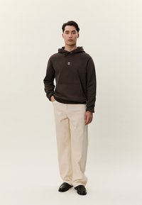 Uomo che indossa una felpa con cappuccio marrone scuro con logo "II", pantaloni beige a gamba larga e scarpe nere, in piedi contro uno sfondo chiaro e uniforme.