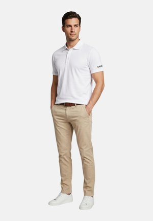 Uomo che indossa una polo bianca a maniche corte, pantaloni beige con cintura marrone e scarpe da ginnastica bianche, in piedi con le mani in tasca.
