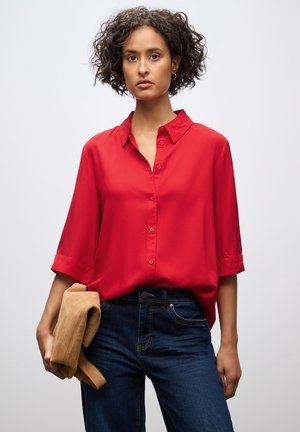 Camicia rossa con bottoni e maniche corte arrotolate, in tessuto leggero, abbinata a jeans blu scuro. Modella che tiene una giacca beige.