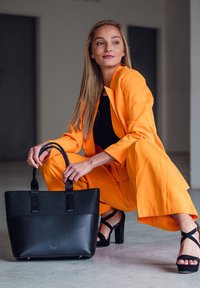 Black leather tote bag with textured finish and two top handles. Model wears an orange suit and black heels, kneeling against a neutral backdrop.