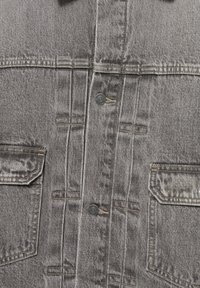 Krupni plan sive traper jakne sprijeda koja prikazuje dva džepa s preklopima, ušivene detalje i crne metalne dugmadi s gravurom brenda.