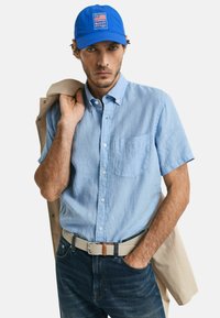 Camisa de manga corta azul claro con un bolsillo en el pecho, combinada con jeans oscuros y un cinturón trenzado. El accesorio incluye una gorra azul con logotipo.