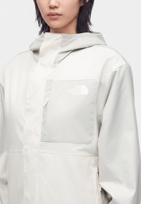 Personne portant une veste beige clair The North Face avec une capuche et une poche poitrine avec le logo de la marque.