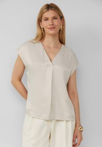 Korte mouwen, lichtbeige blouse van een gladde stof met een V-hals en een plooidetail aan de voorkant. Draagt met crèmekleurige broek en gouden sieraden.