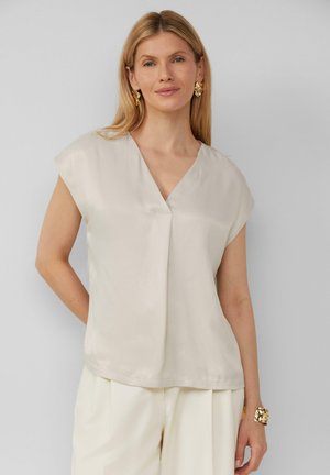 Korte mouwen, lichtbeige blouse van een gladde stof met een V-hals en een plooidetail aan de voorkant. Draagt met crèmekleurige broek en gouden sieraden.