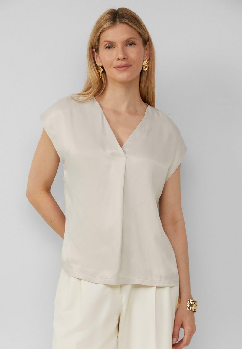 Korte mouwen, lichtbeige blouse van een gladde stof met een V-hals en een plooidetail aan de voorkant. Draagt met crèmekleurige broek en gouden sieraden.
