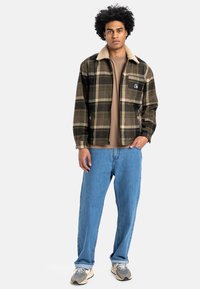 Rudolfærklæde i grøn, sort og beige med en pelsforet krave; parret med en tan skjorte og løse blå jeans. Snickers med grå detaljer.