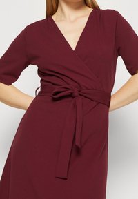 Un vestido envolvente burdeos hecho de una tela suave con mangas cortas, que presenta un cinturón atado en la cintura y un escote en V profundo.
