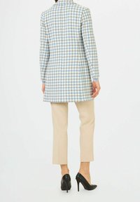 Manteau long à motif pied-de-poule bleu clair et blanc, coupe ajustée, avec un col à revers et des manches longues, associé à un pantalon crème et des talons noirs.