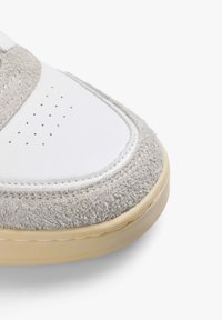 Zapatilla blanca con acentos de ante gris. Forma redondeada, puntera de cuero suave, perforaciones para transpirabilidad y suela de goma de color beige claro.