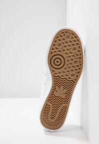 Vitt tygsko med gummisula som har ett texturerat brunt mönster. Tre cirkulära detaljer och en logotyp på sulan.