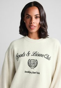 Cremefarvet sweater med ribbestrikket halsudskæring, prydet med sort broderet tekst "Sports & Leisure Club" og et cirkulært emblem med teksten "Est. 1989."