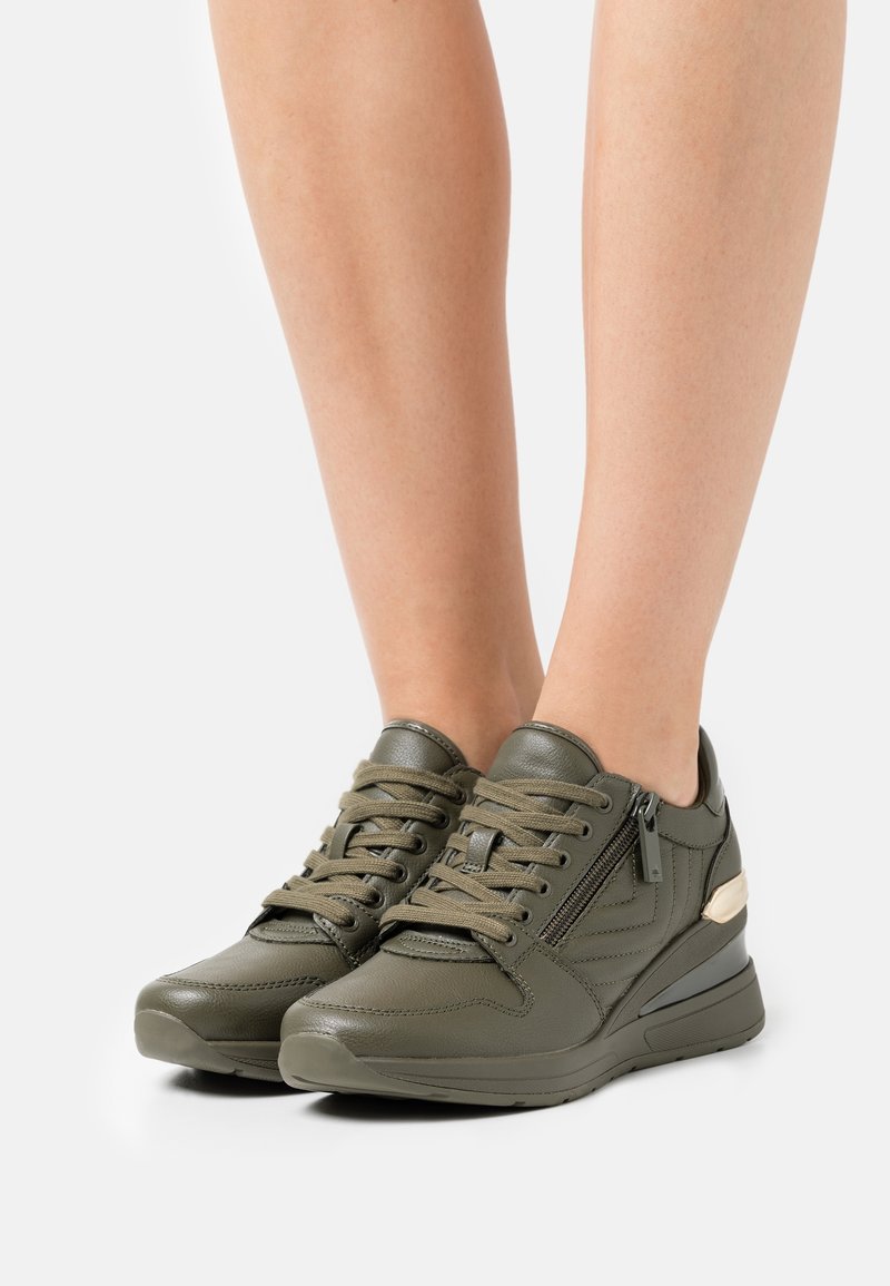 Pieds portant des baskets en cuir vert olive avec des lacets et une fermeture éclair latérale, posés sur une surface blanche.
