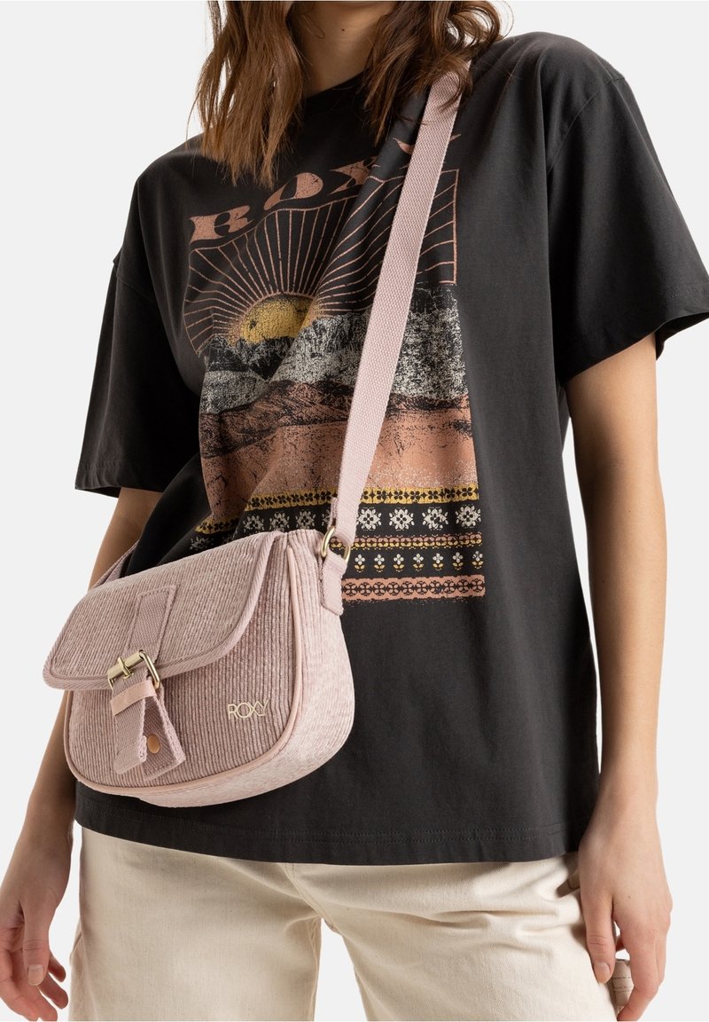 Růžová kordurová crossbody taška s klopou a zlatými kovovými doplňky, má strukturovaný tvar a detail s logem, nošená s tričkem s potiskem.