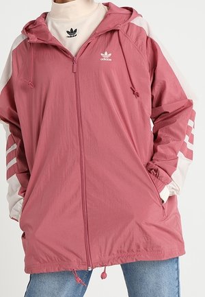 Osoba ubrana w różową kurtkę Adidas z kapturem, z białymi i różowymi paskowanymi rękawami, białym golfem oraz niebieskimi dżinsami.